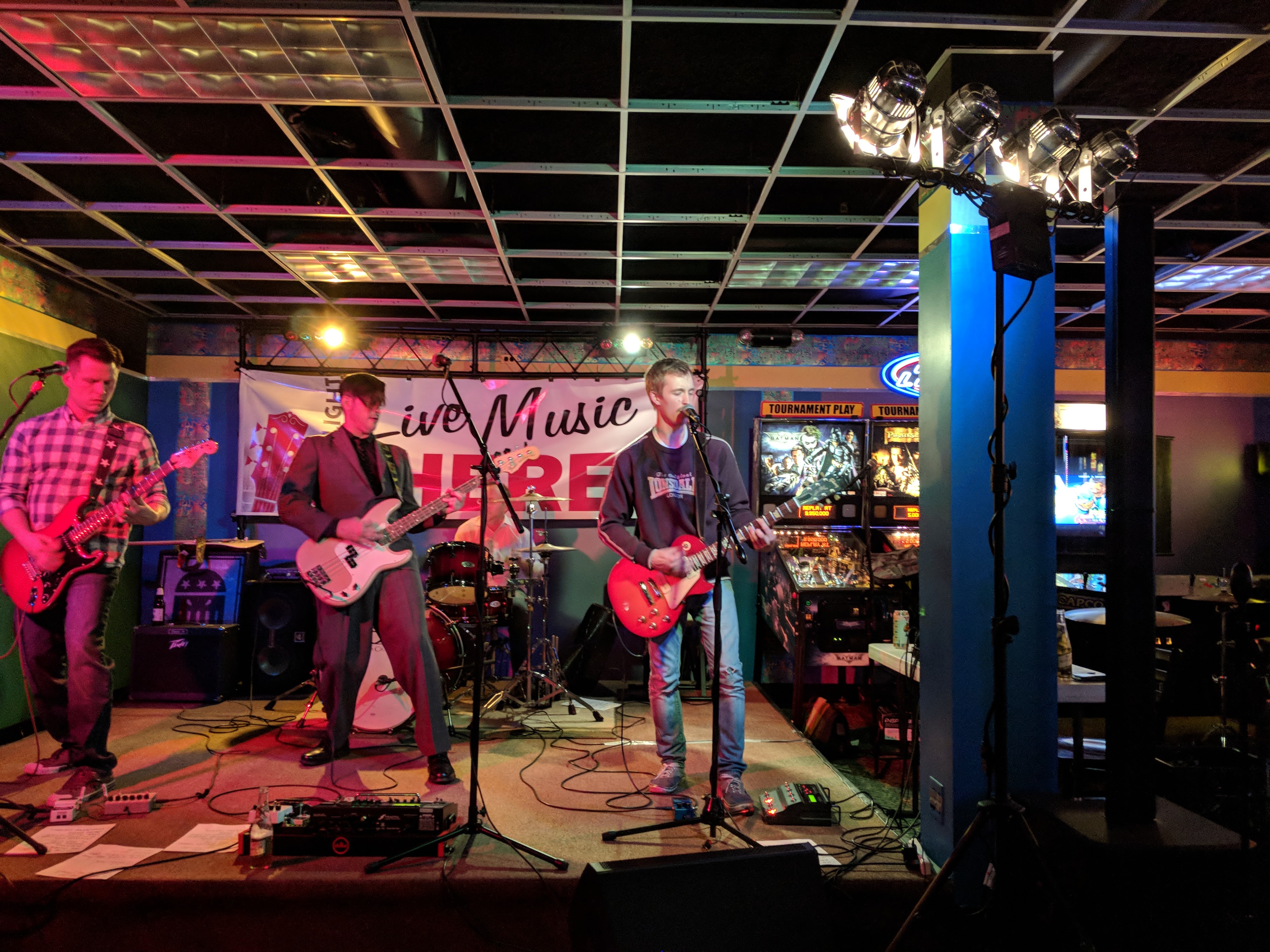 A four-piece band performs on a stage with a 'Live Music HERE' banner and pinball machines in the background.