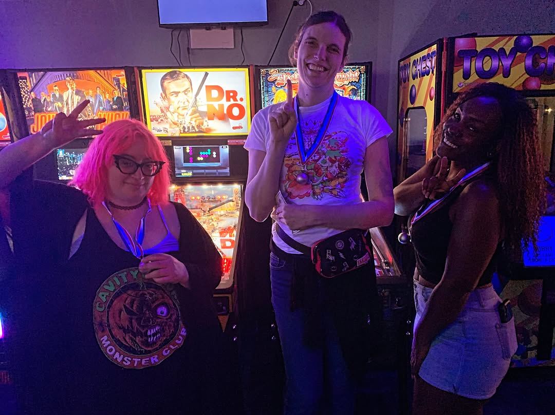 Three people smiling and wearing medals in front of pinball machines in an arcade.