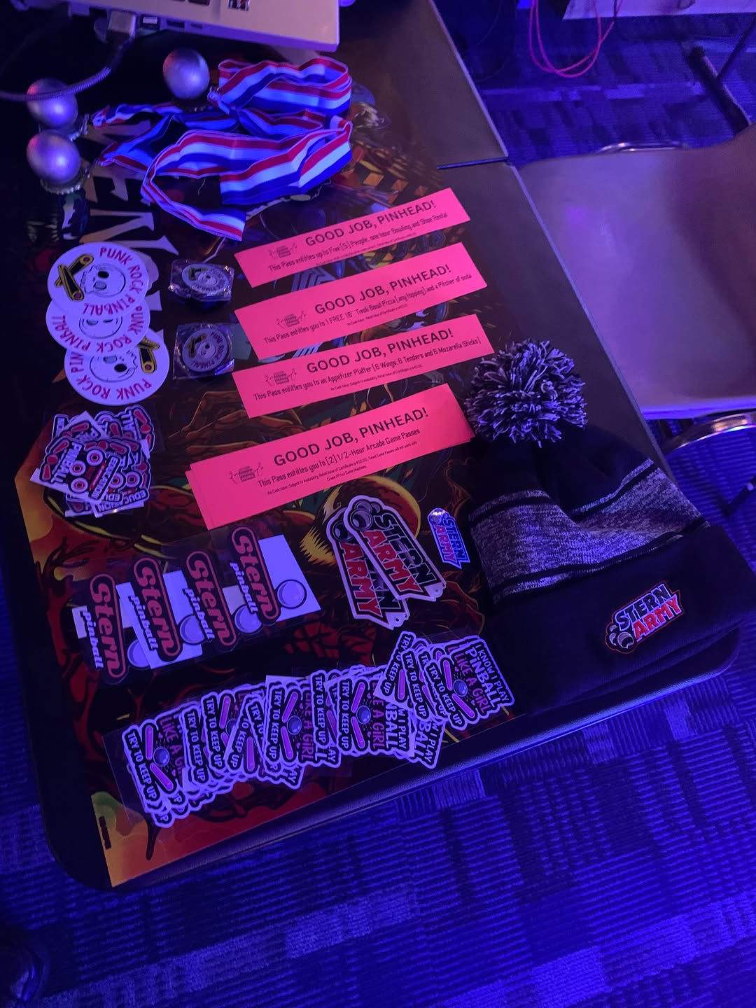 Pinball stickers, prize vouchers, a beanie, and medals arranged on a table under purple cosmic lighting.