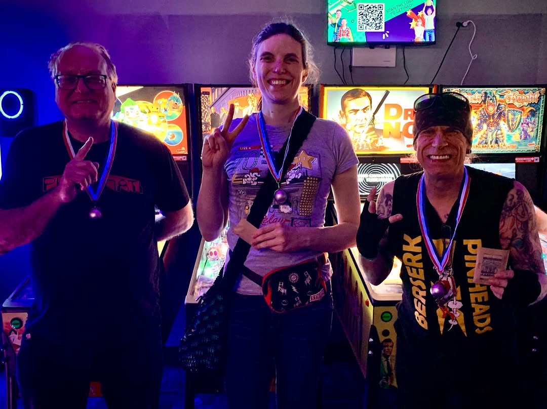 Three tournament winners wearing medals pose in front of pinball machines in a brightly lit arcade.