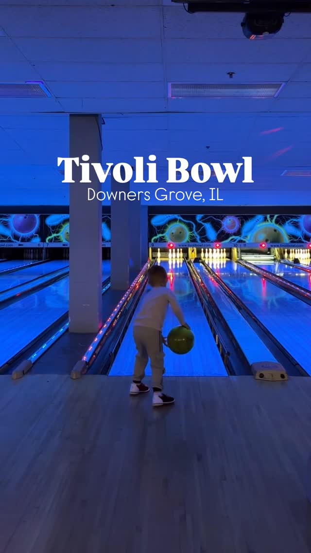 A young child stands at the bowling lane foul line holding a green bowling ball under blue cosmic lights.