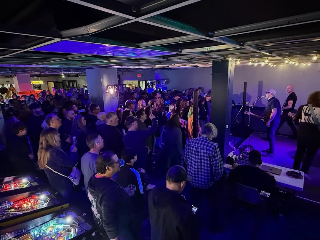 A large crowd watches a live band perform in a lounge area next to several pinball machines.