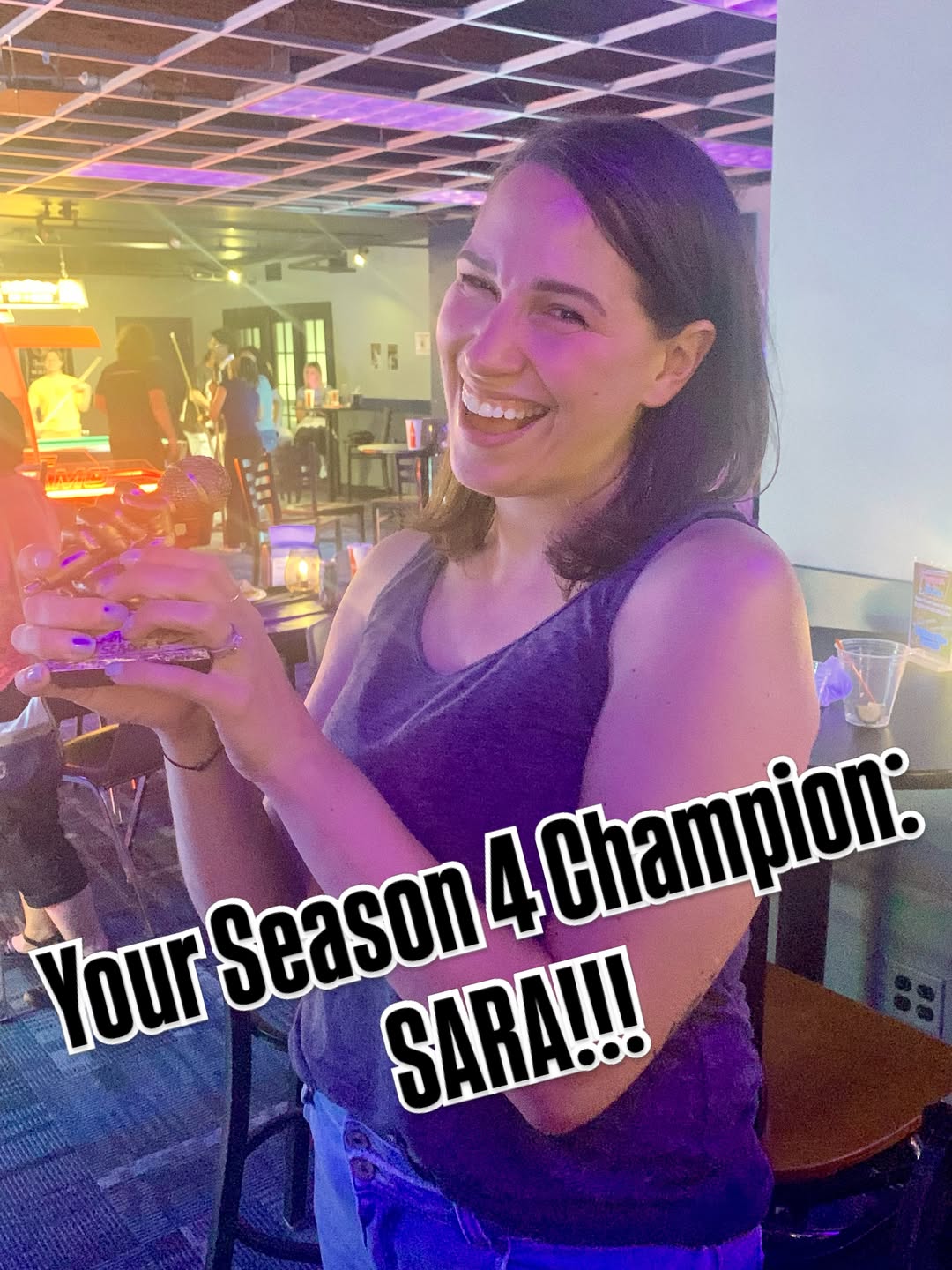 A woman smiles and laughs while holding a microphone trophy in a dimly lit lounge with pool tables in the background.