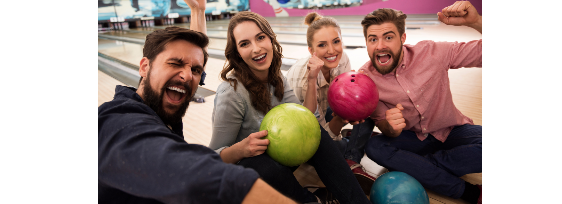 Four friends cheer and hold colorful bowling balls while sitting together on a bowling lane.