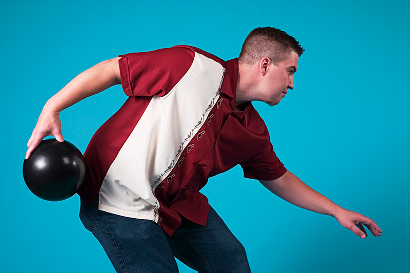A man in a red and white bowling shirt leans forward to throw a black bowling ball against a solid blue background.