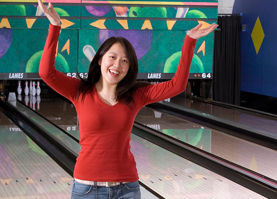 A woman in a red shirt celebrates with her arms raised in front of bowling lanes.