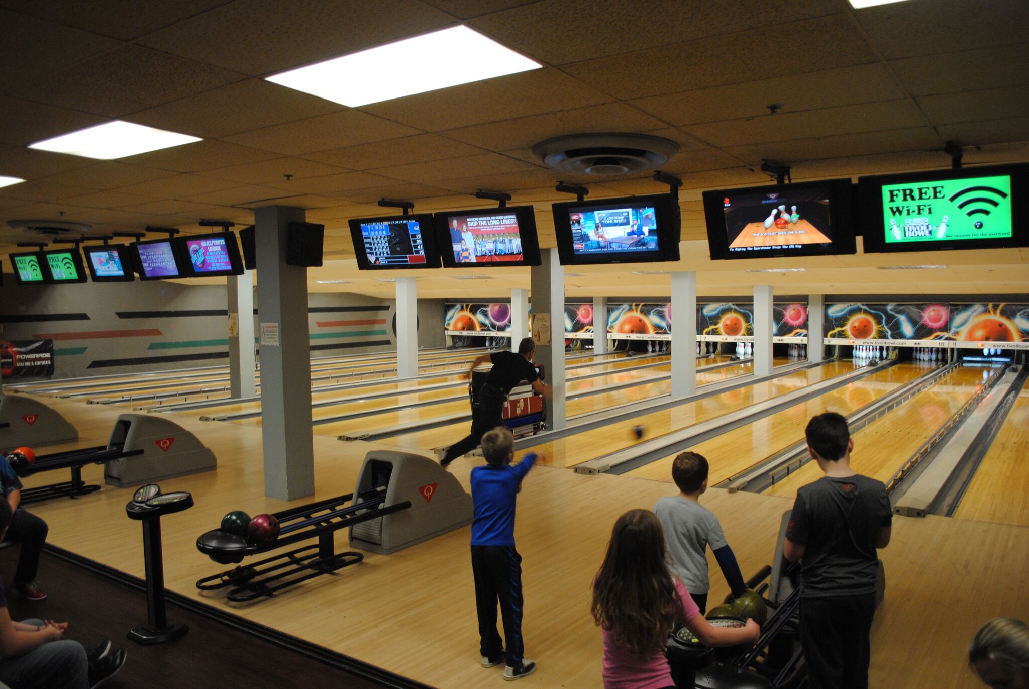 A group of children and an adult bowling on well-lit wooden lanes under overhead score monitors.