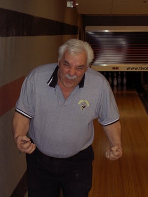 An older man in a grey Tivoli Bowl polo shirt celebrating on a bowling lane.