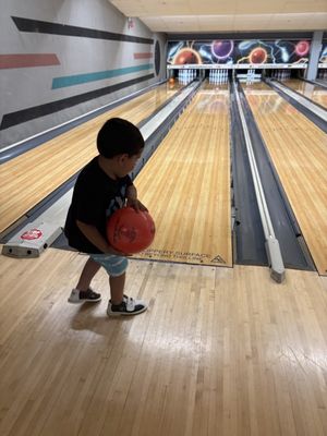 A young boy in a black shirt holds a red bowling ball at the start of a wooden bowling lane.