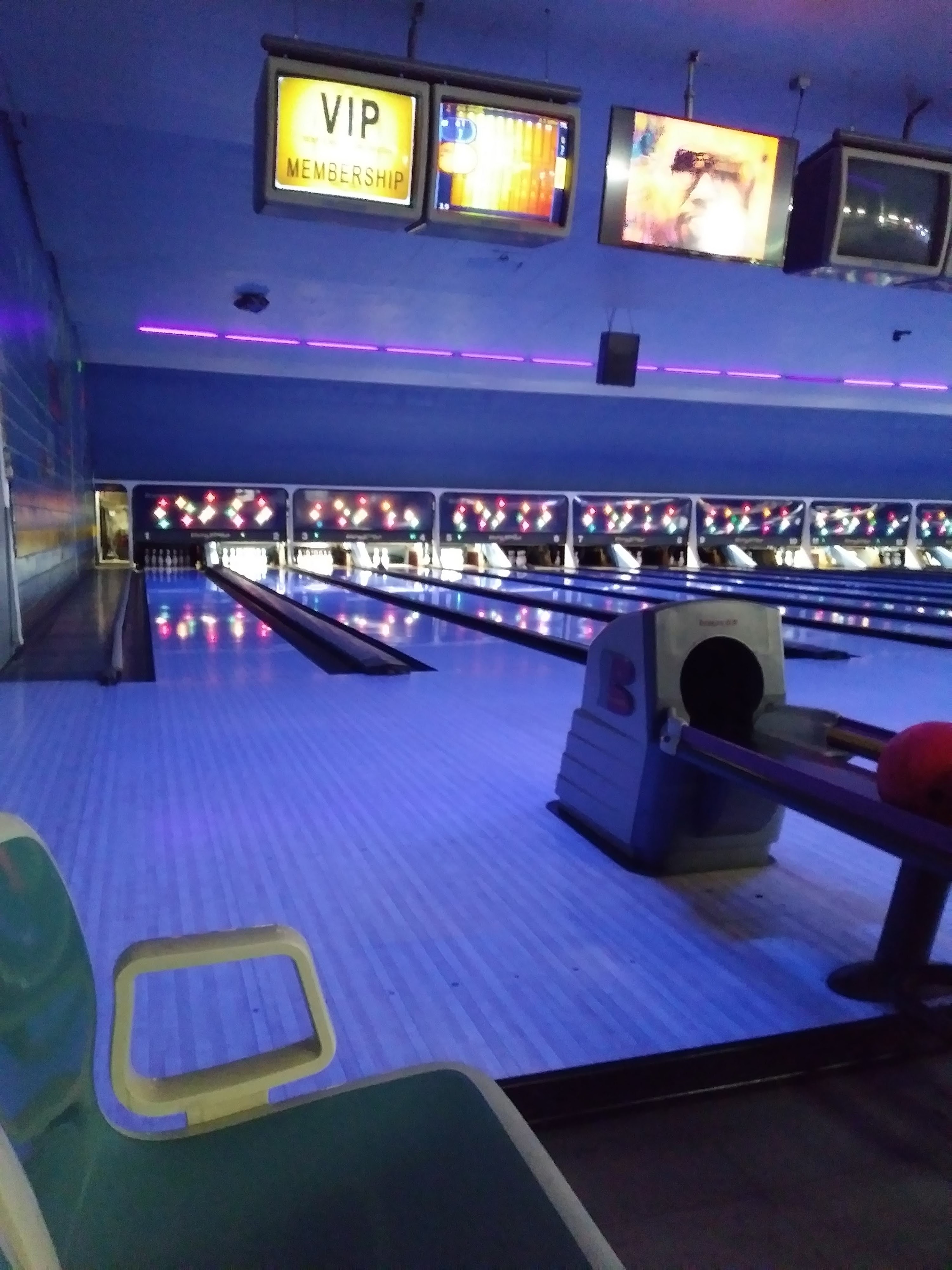 A bowling alley at night with blue glow-in-the-dark lighting, wooden lanes, and overhead monitors.