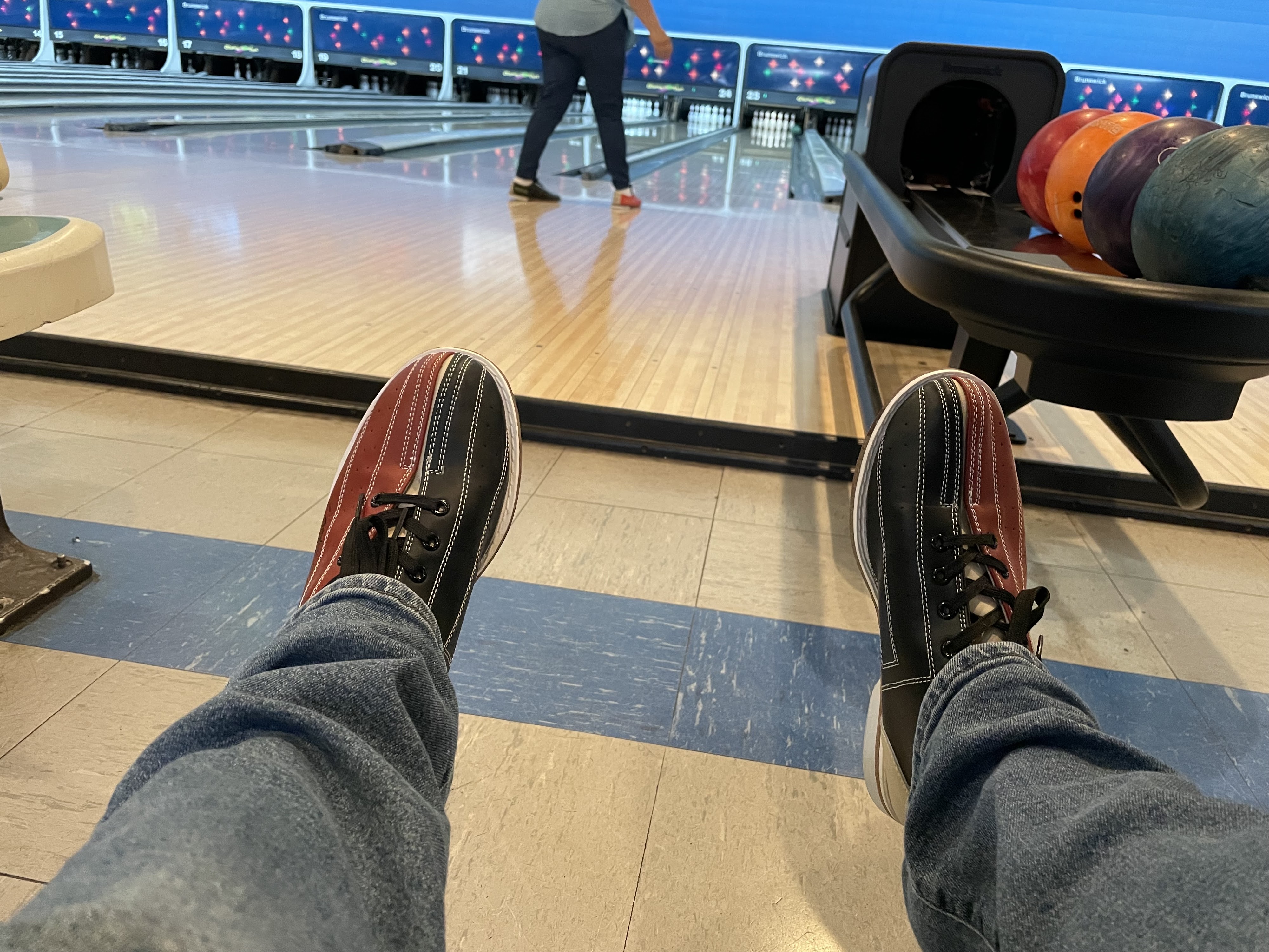 A first-person view of red and black rental bowling shoes with bowling lanes and a ball return in the background.