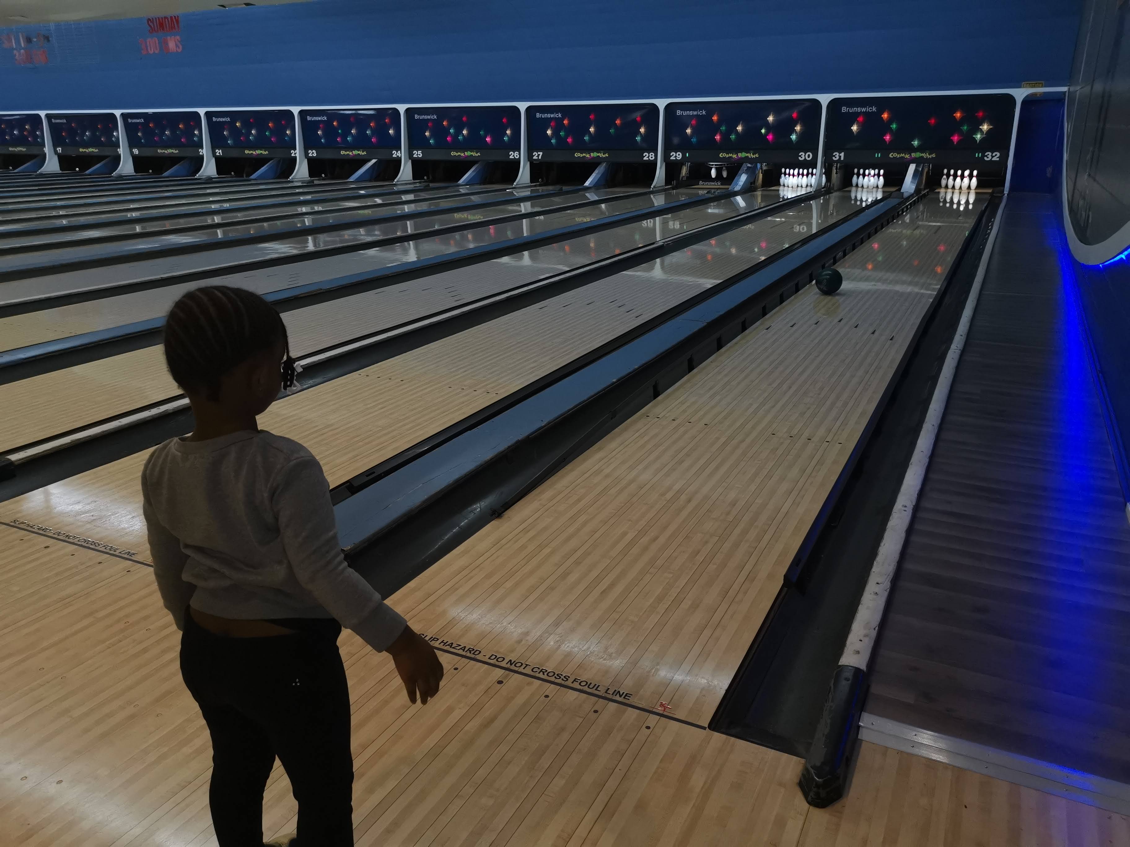 A young child stands behind the foul line watching a bowling ball travel down a wooden lane toward the pins.