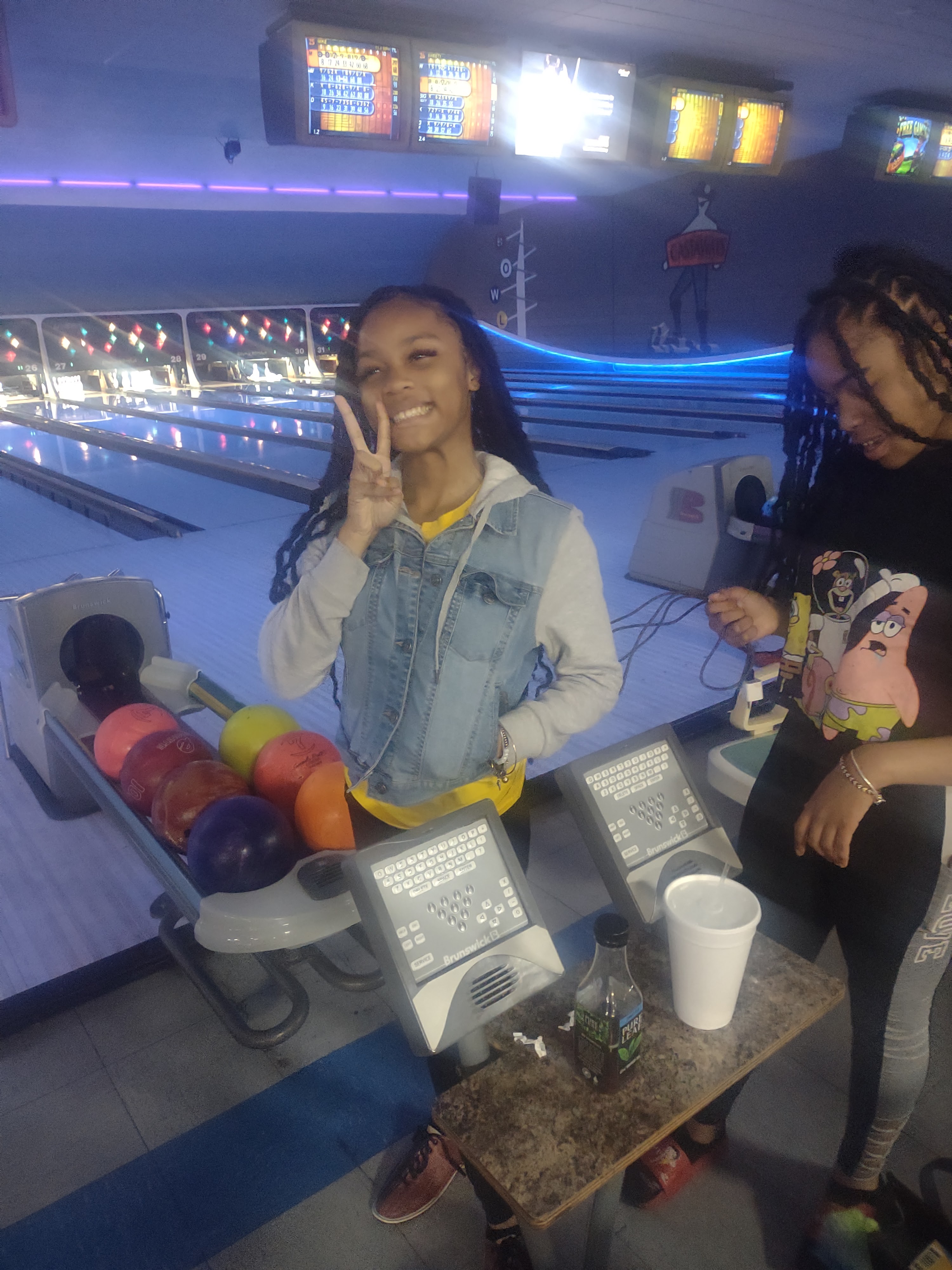 A smiling young woman gives a peace sign while standing by a bowling ball return under neon lights.