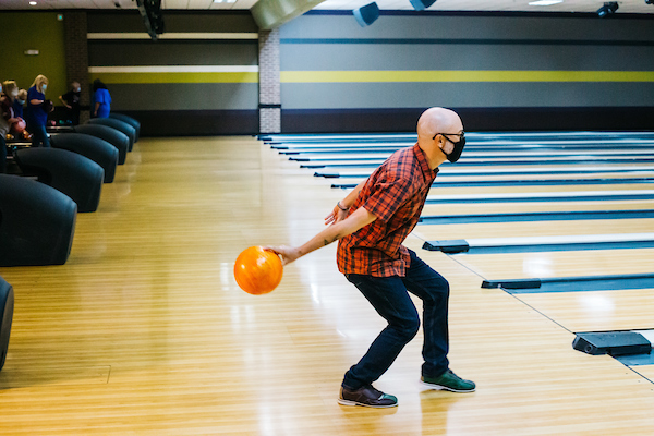 A man in a plaid shirt and face mask prepares to roll an orange bowling ball down a lane.