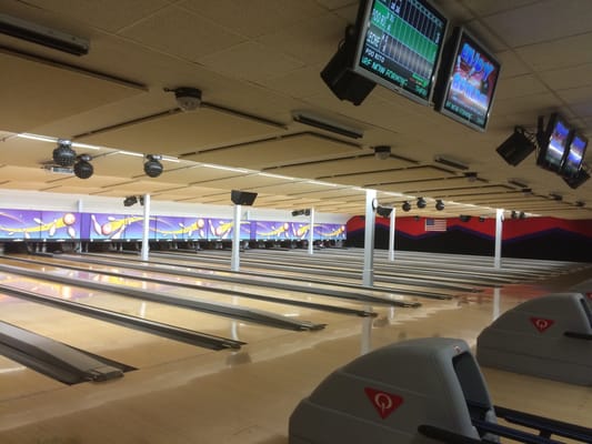 A view down a long row of polished bowling lanes with overhead scoring monitors and bright ceiling lighting.