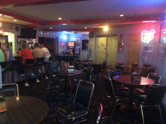 A dimly lit bowling alley lounge with patrons at the bar, circular tables, and a pinball machine in the background.