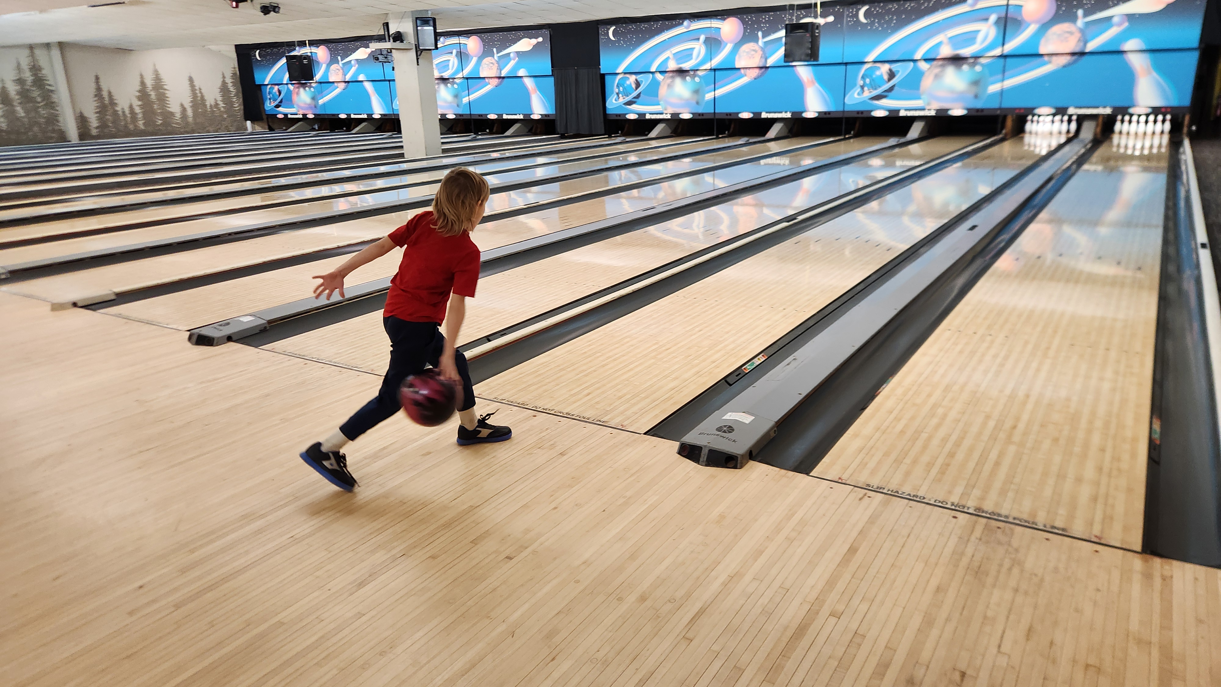 A young bowler in a red shirt releases a bowling ball on a wooden lane at a large bowling alley.