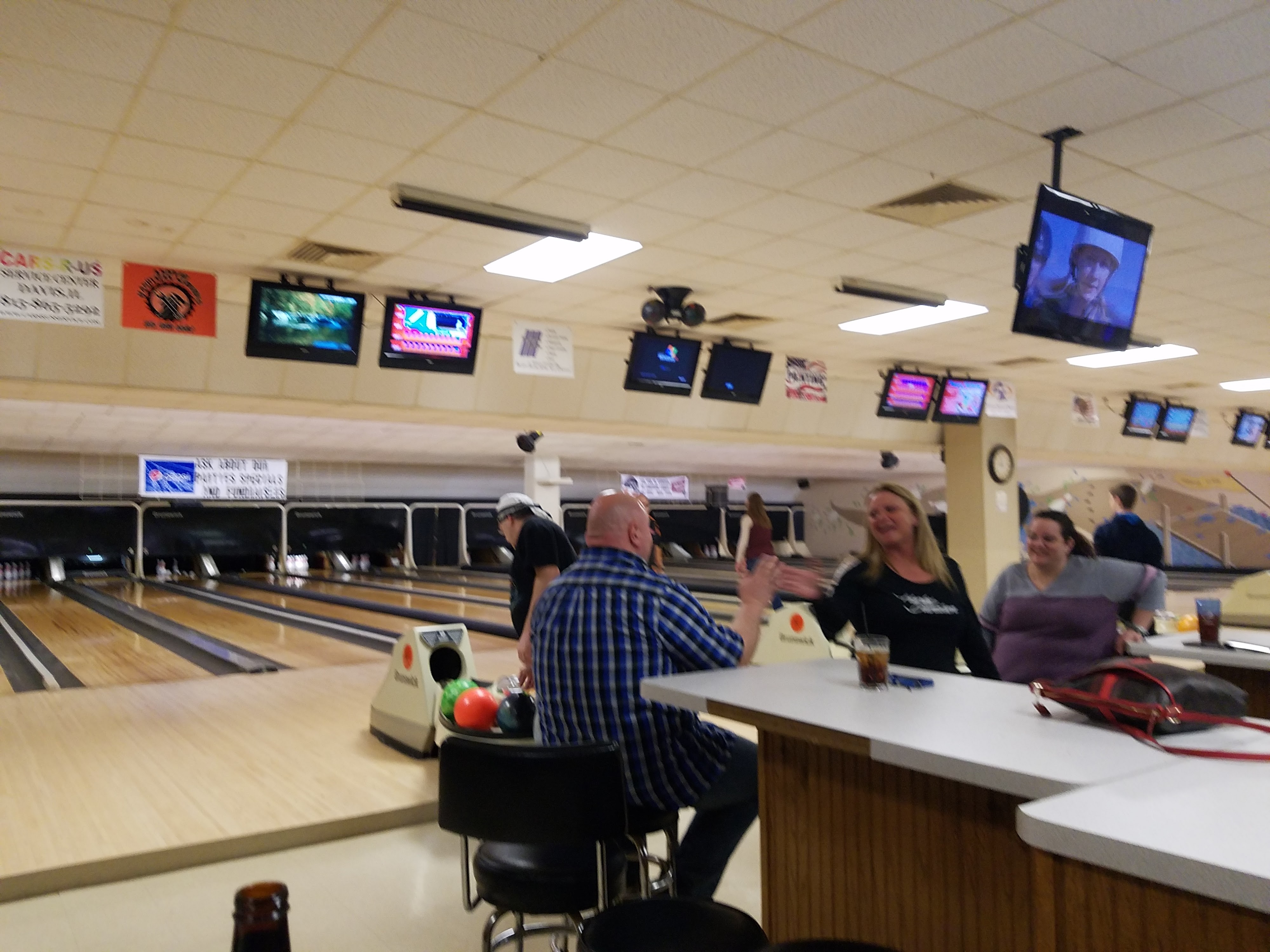 A man in a plaid shirt and a woman in a black shirt high-fiving at a table in a bowling alley.
