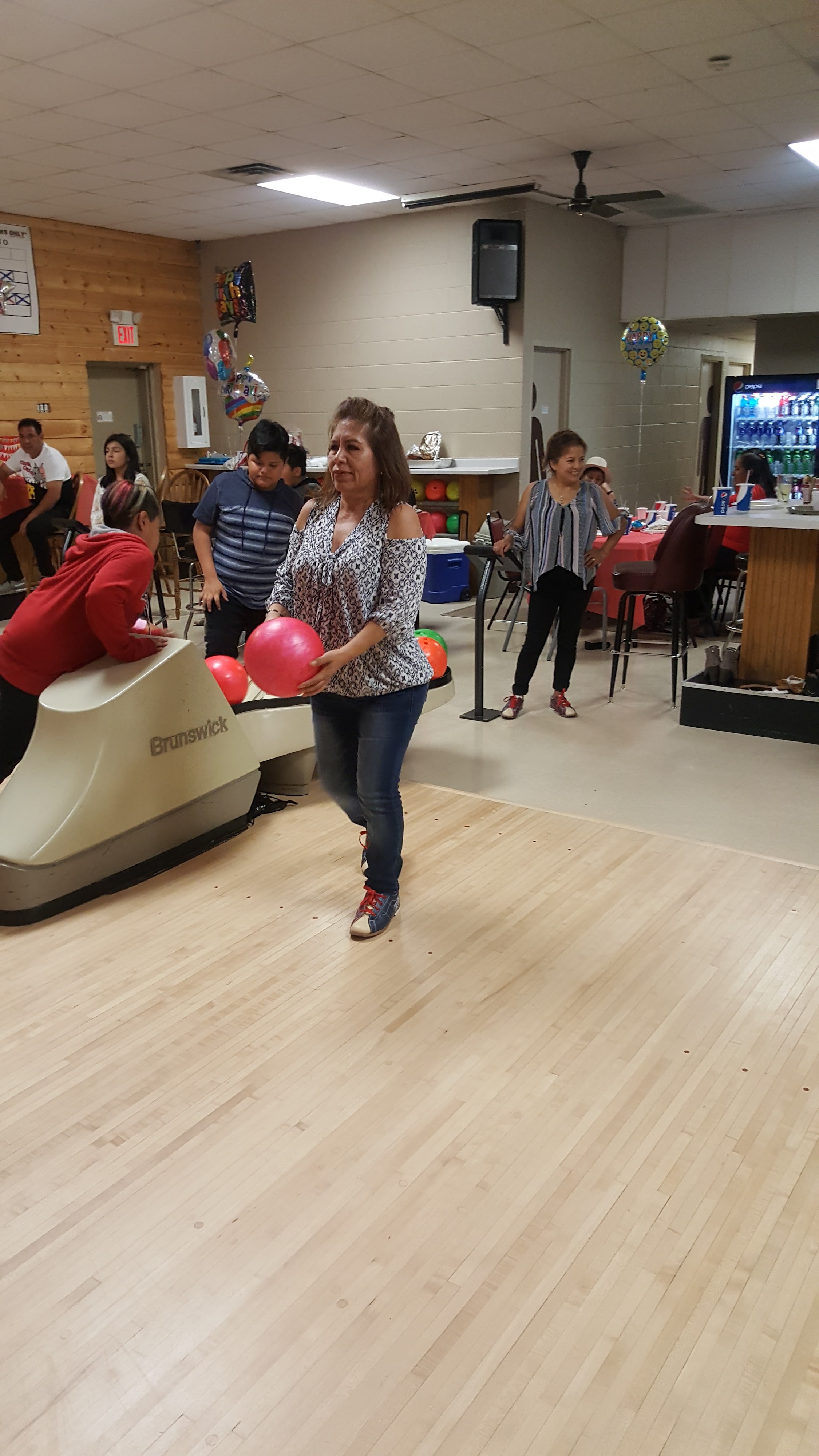 A woman in a patterned shirt holds a pink bowling ball while standing on a wooden bowling lane.