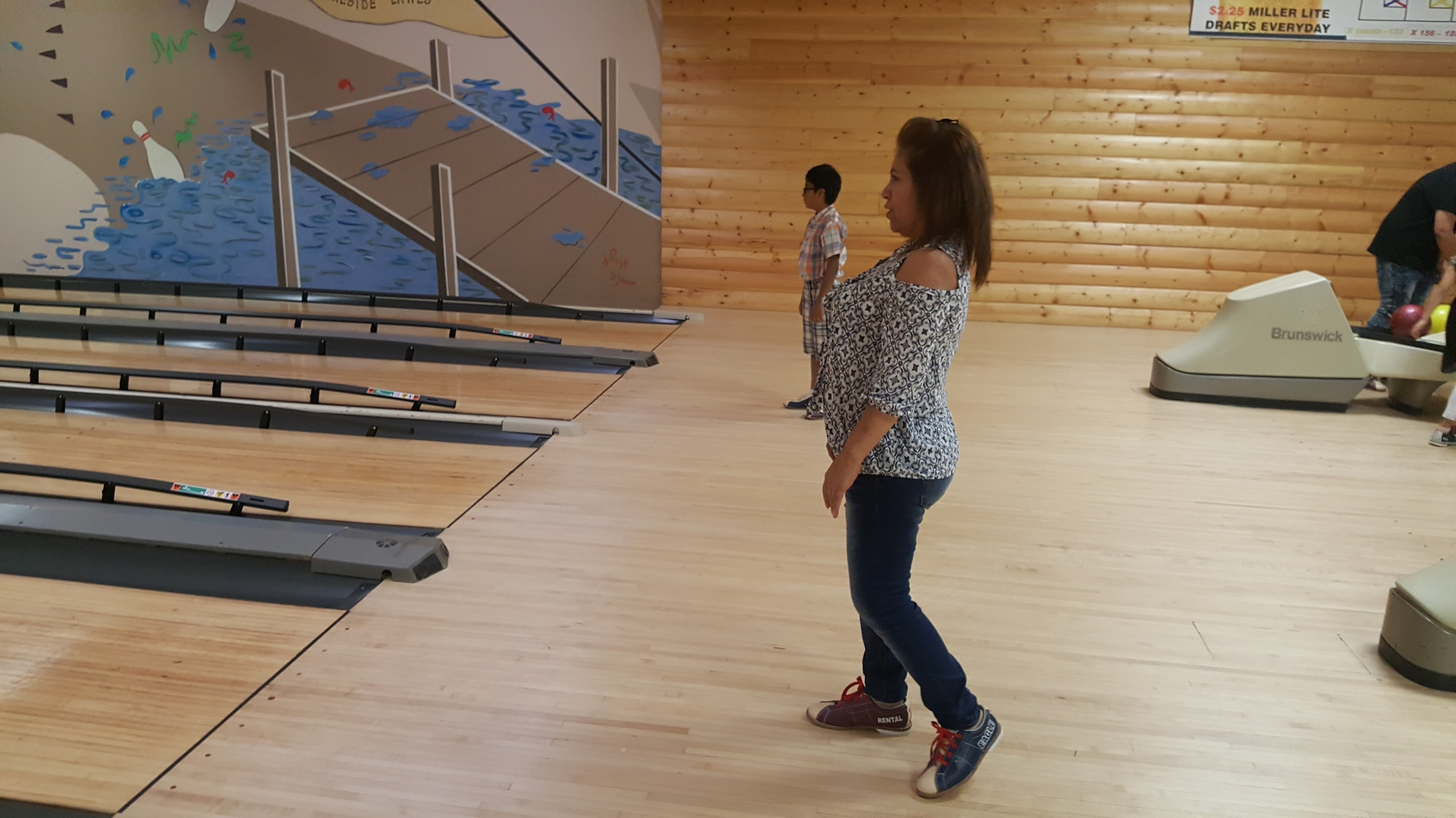 A woman and a child stand on the wooden approach area of a bowling alley with a lake mural in the background.