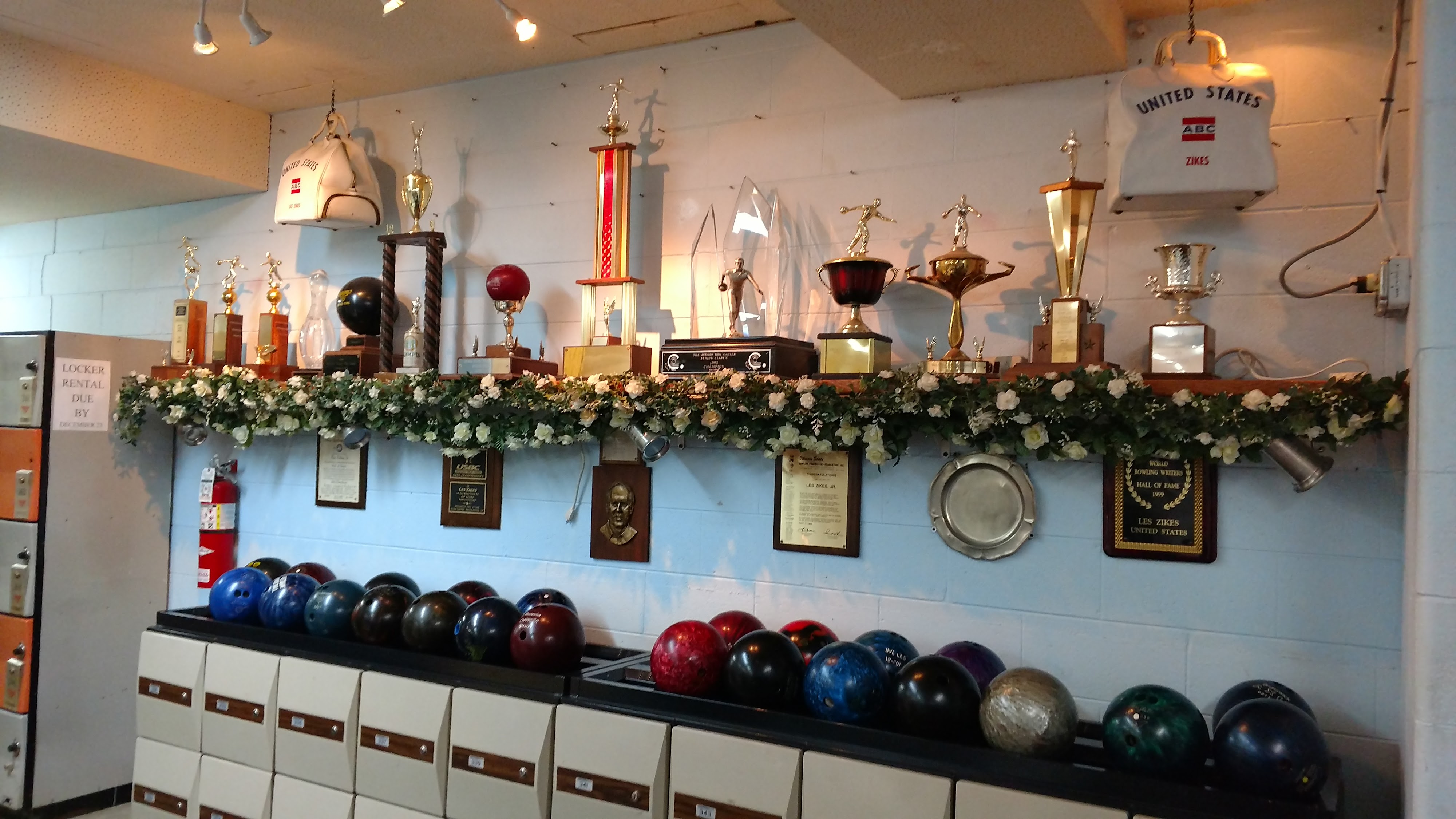 A wall display of historical bowling trophies and plaques above a rack of colorful house bowling balls.