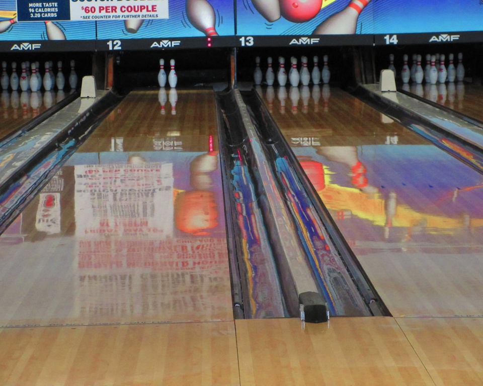 A low-angle view of three polished bowling lanes with pins at the end and colorful reflections on the surface.
