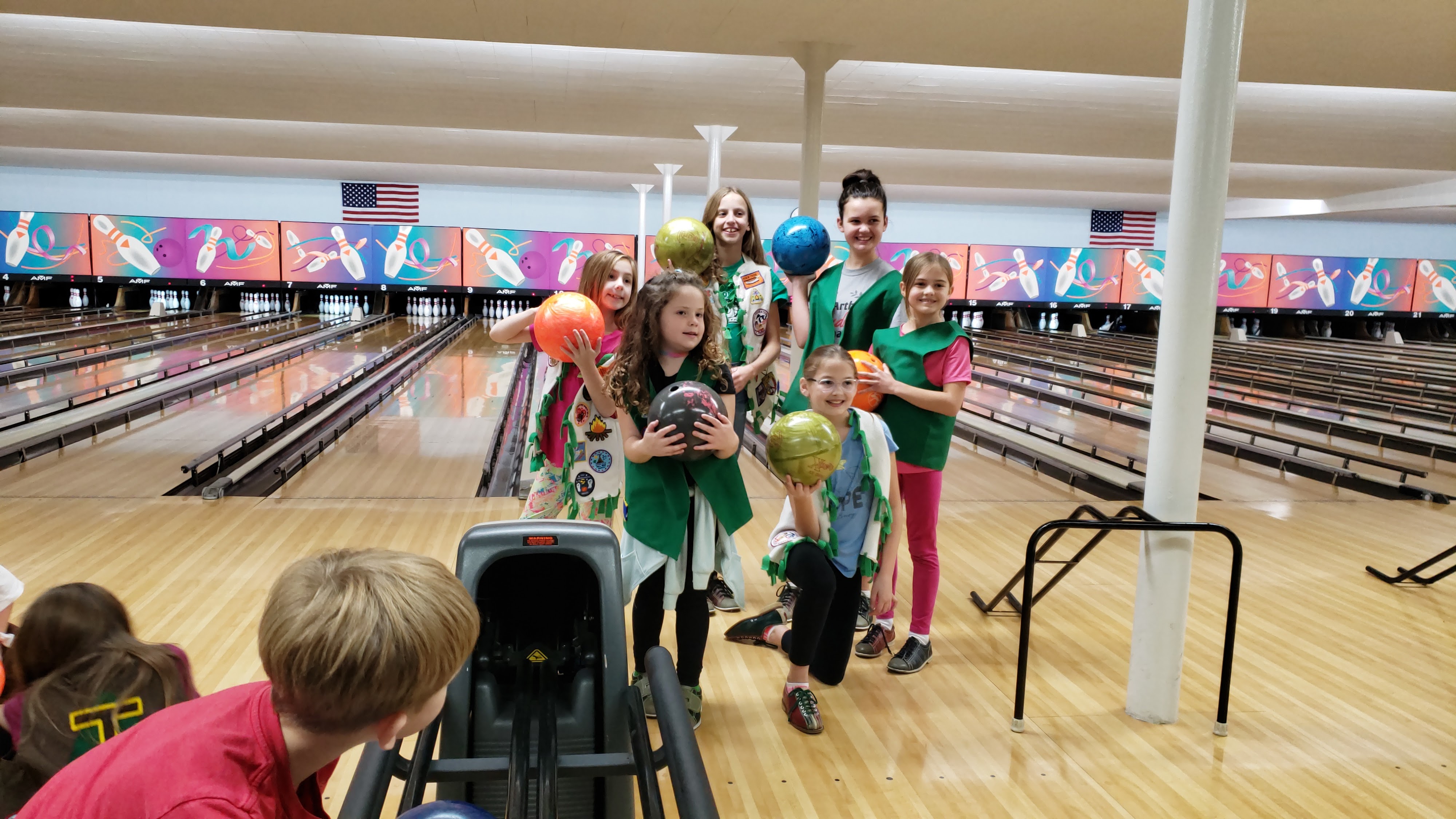 A group of young girls in green scout vests holding colorful bowling balls and smiling on the lanes.