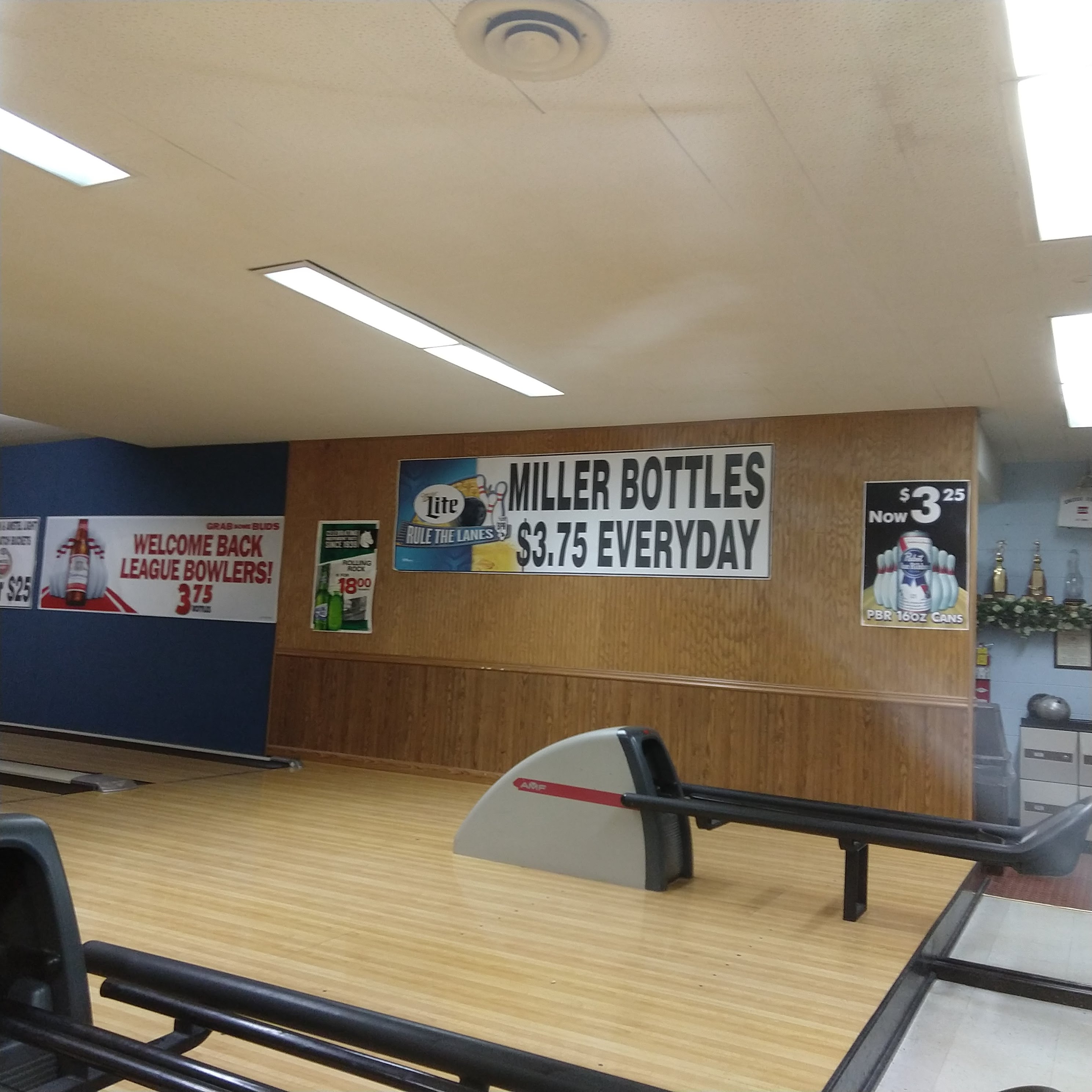 A bowling ball return on a wood floor in front of a wall with league welcome banners and beer special signs.