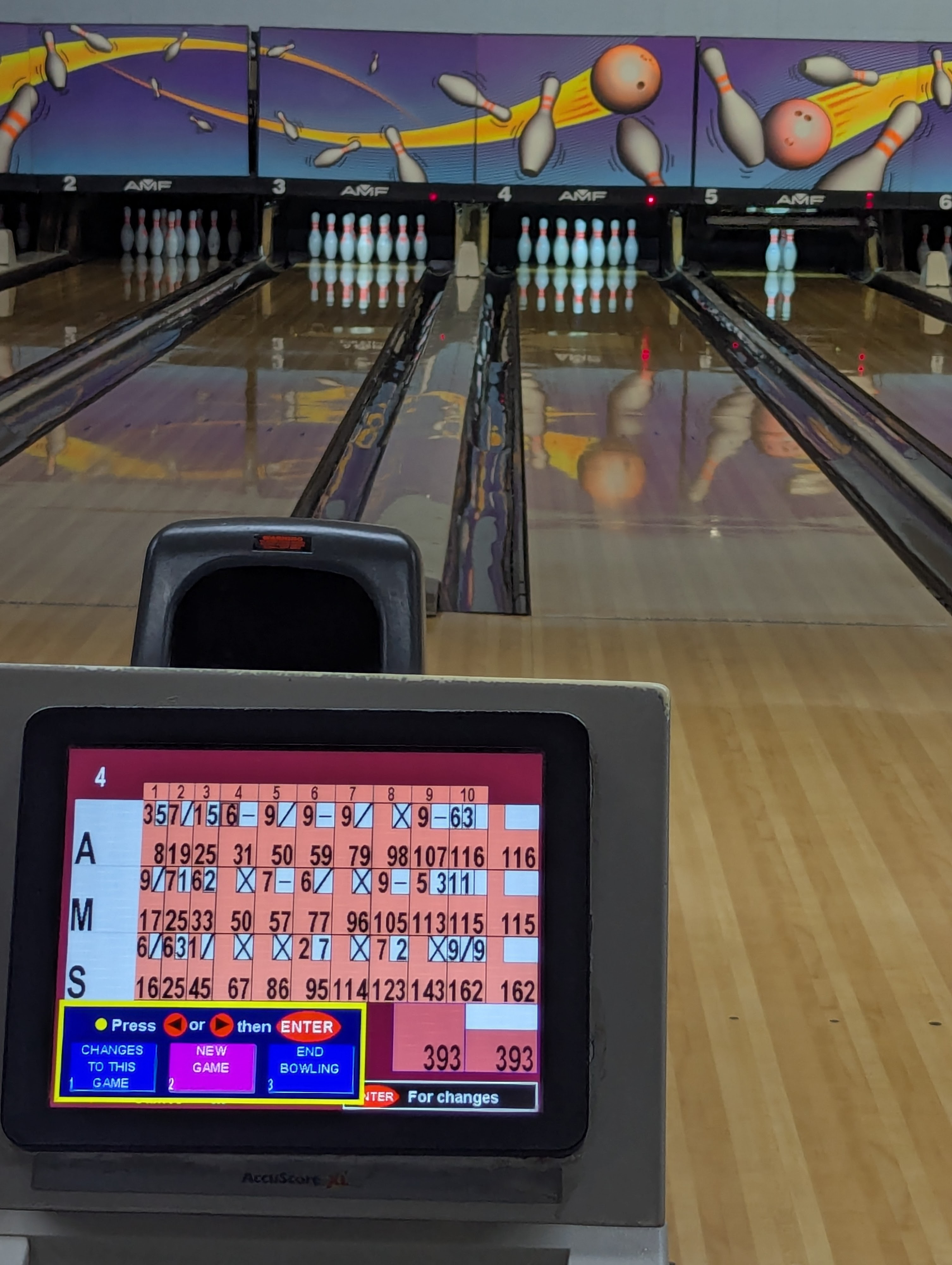 A digital scoring monitor displaying game results in front of polished bowling lanes with pins set.