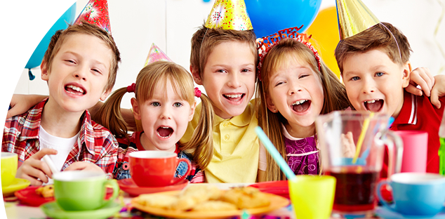 Five smiling children wearing colorful party hats sit at a table with balloons and snacks.
