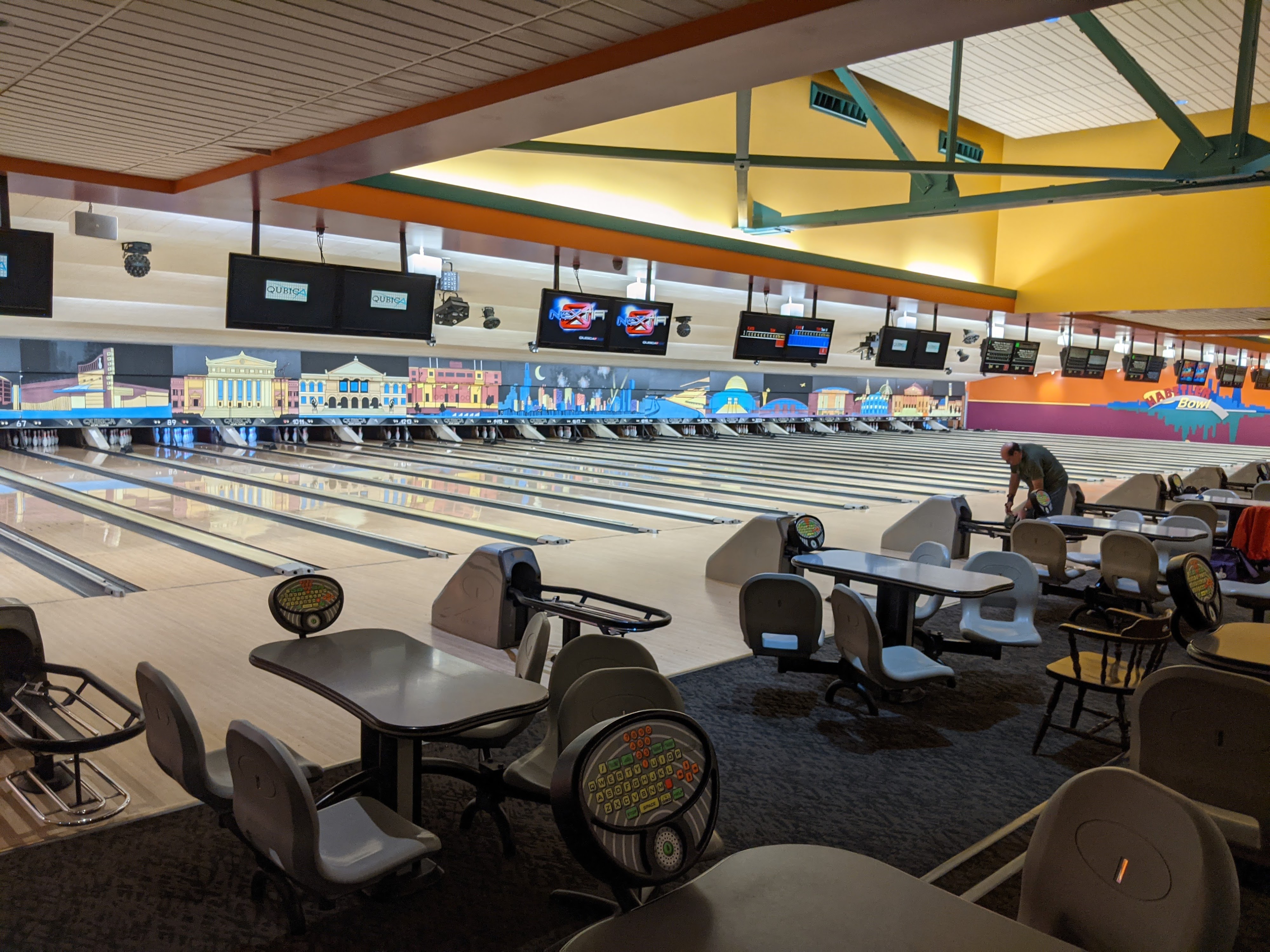 Wide view of a brightly lit bowling center with many lanes, modern overhead scoring monitors, and a colorful mural.