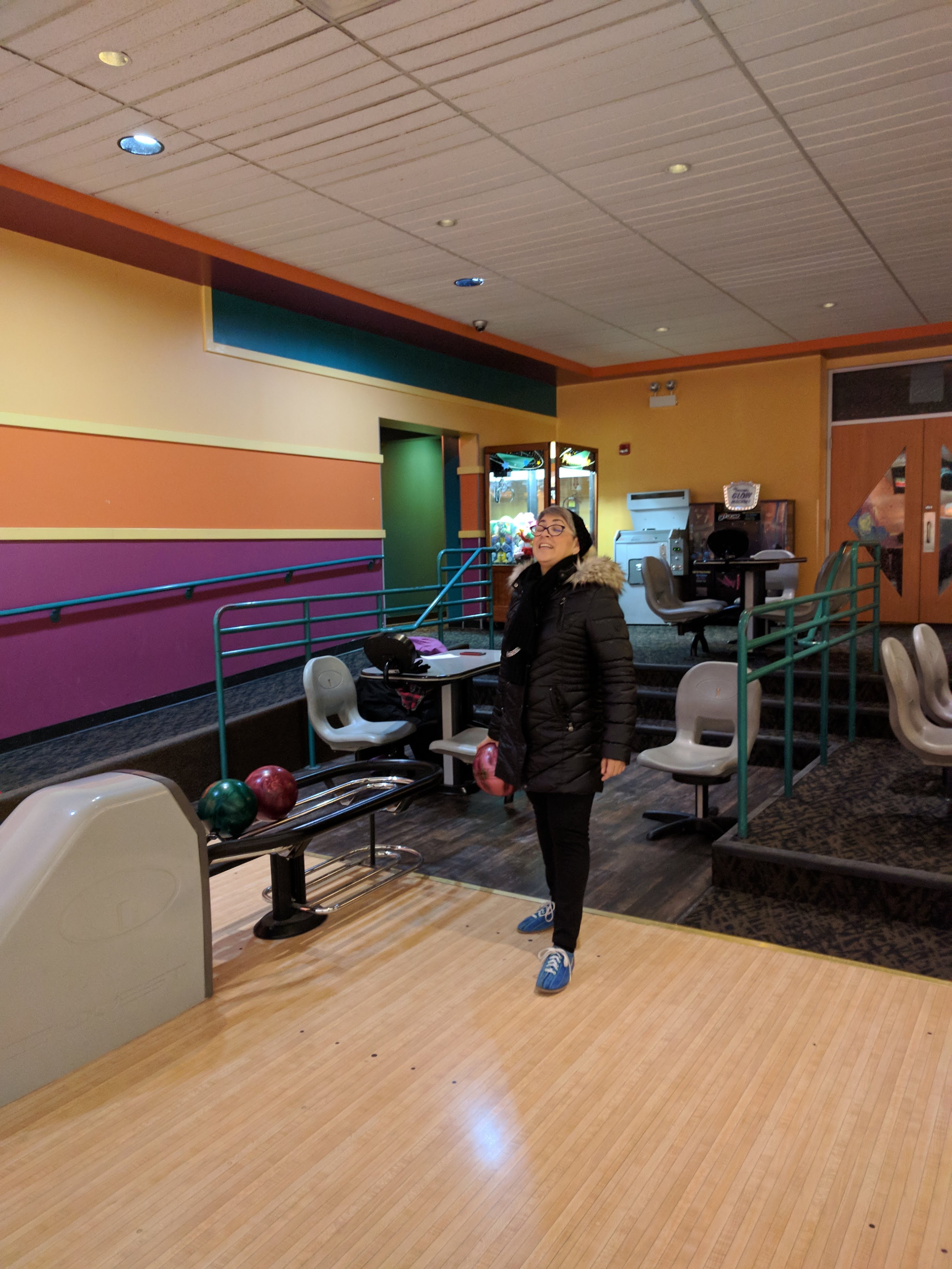A woman in a winter coat holds a bowling ball and smiles on a brightly colored bowling lane.