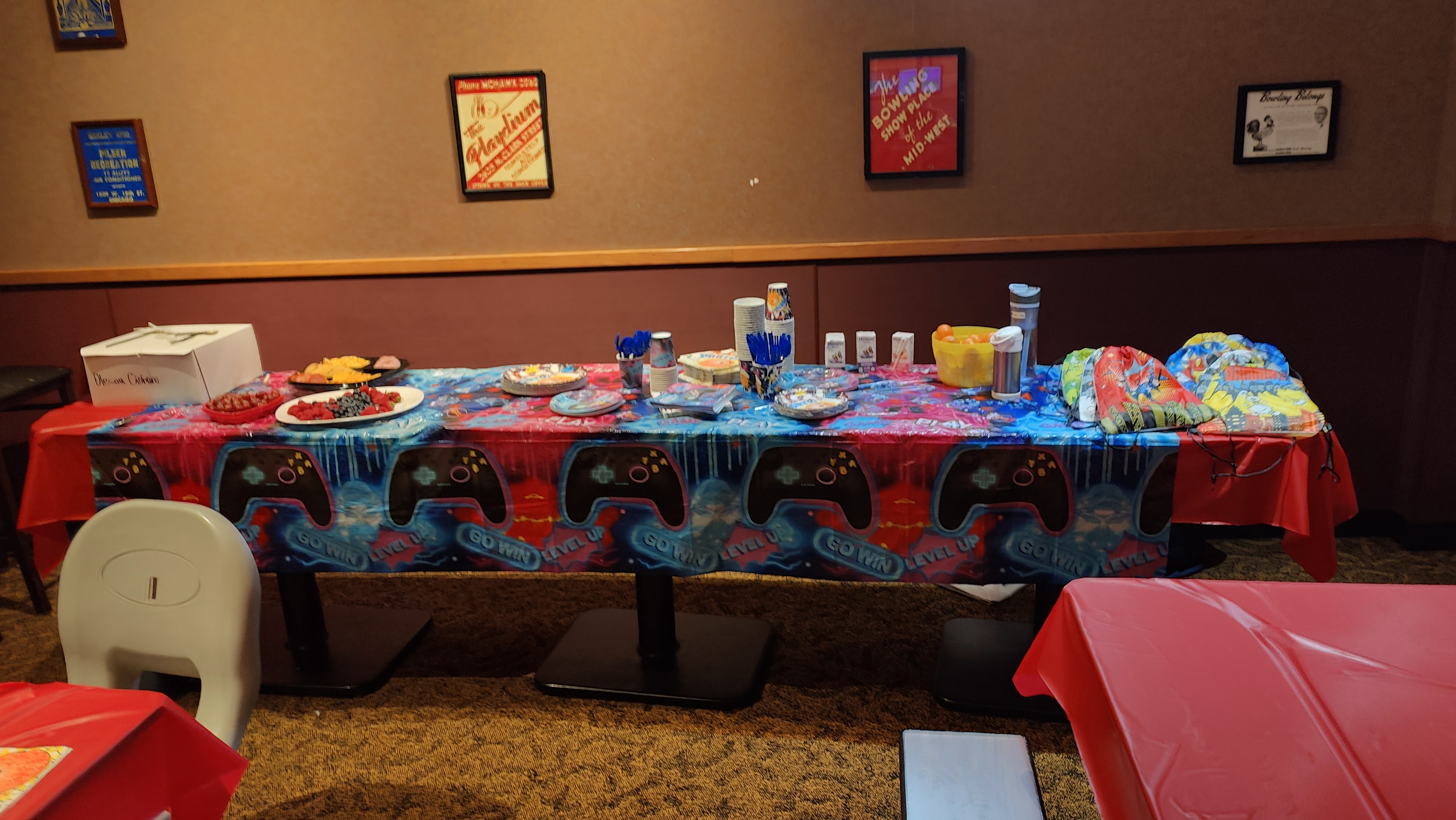 A party table with a gaming-themed tablecloth, fruit platters, and party favors in a bowling center.