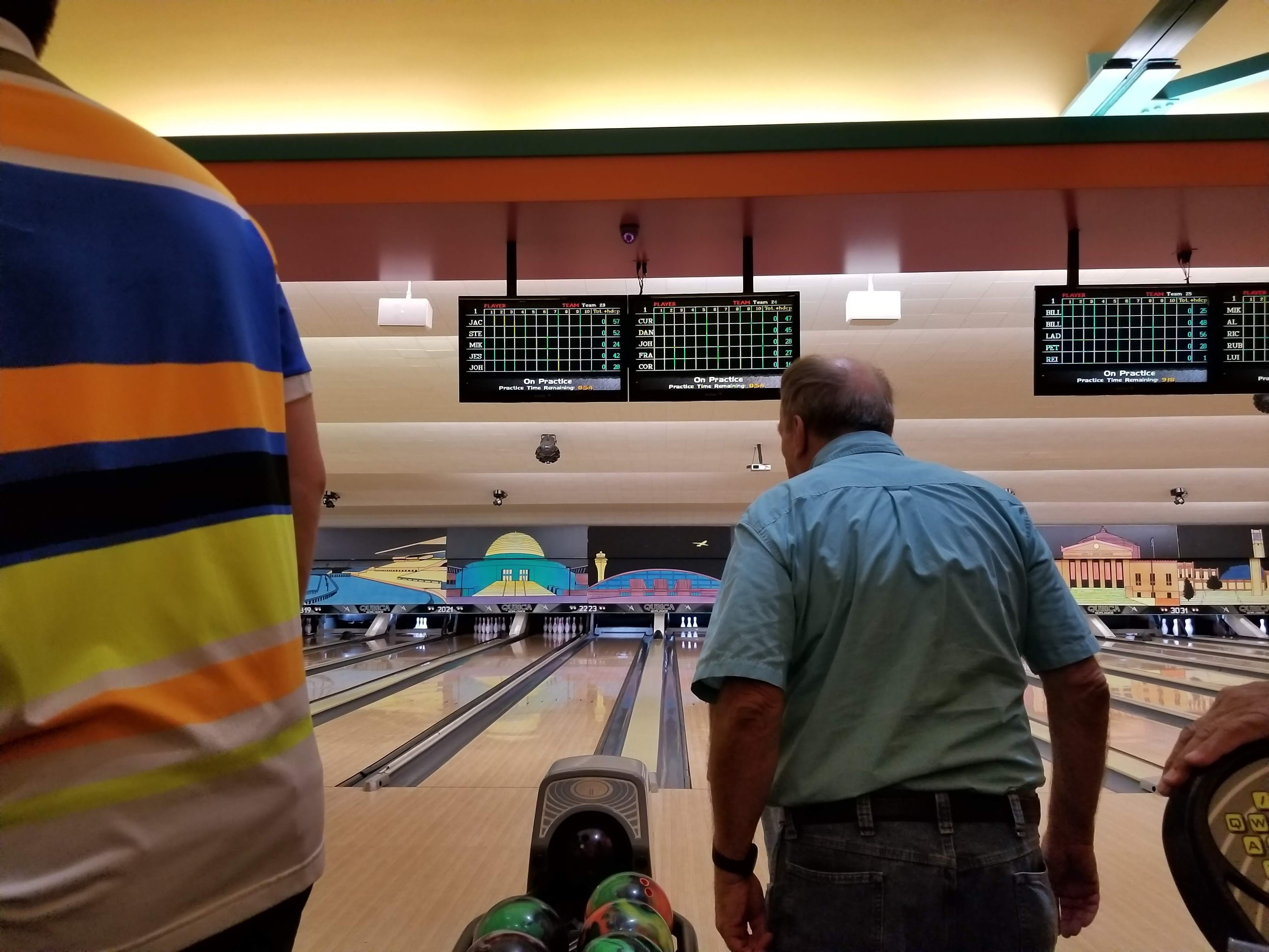 Rear view of two bowlers at the approach with overhead monitors displaying scores and player names.