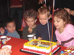 A group of children gather around a lit birthday cake on a red table.