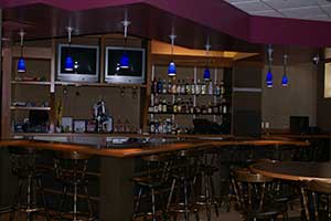 A wooden bar area with blue pendant lights, bar stools, and two televisions.