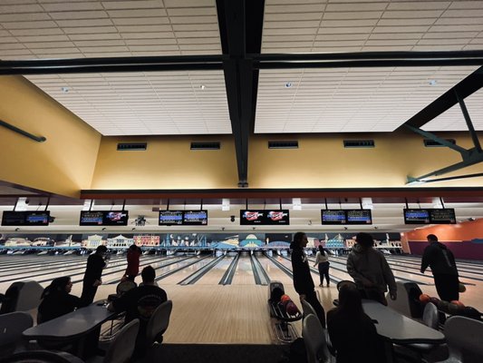 Wide view of people bowling at an indoor center with scoring monitors and a city mural on the back wall.