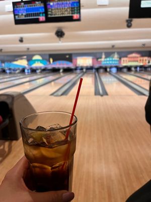 A hand holding a drink with a red straw in front of active bowling lanes and scoring monitors.