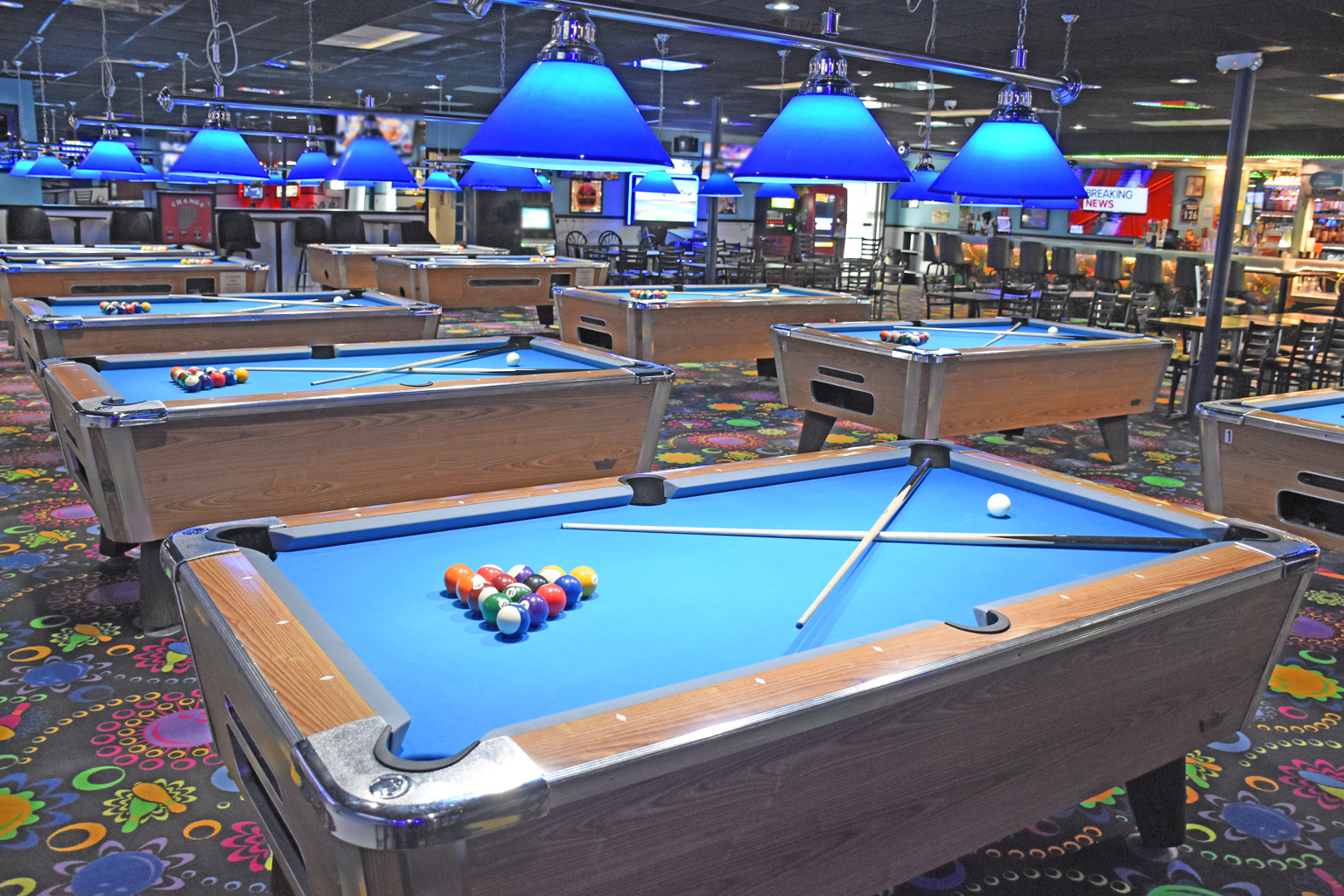 Several pool tables with blue felt under bright blue hanging lamps in a recreation center.