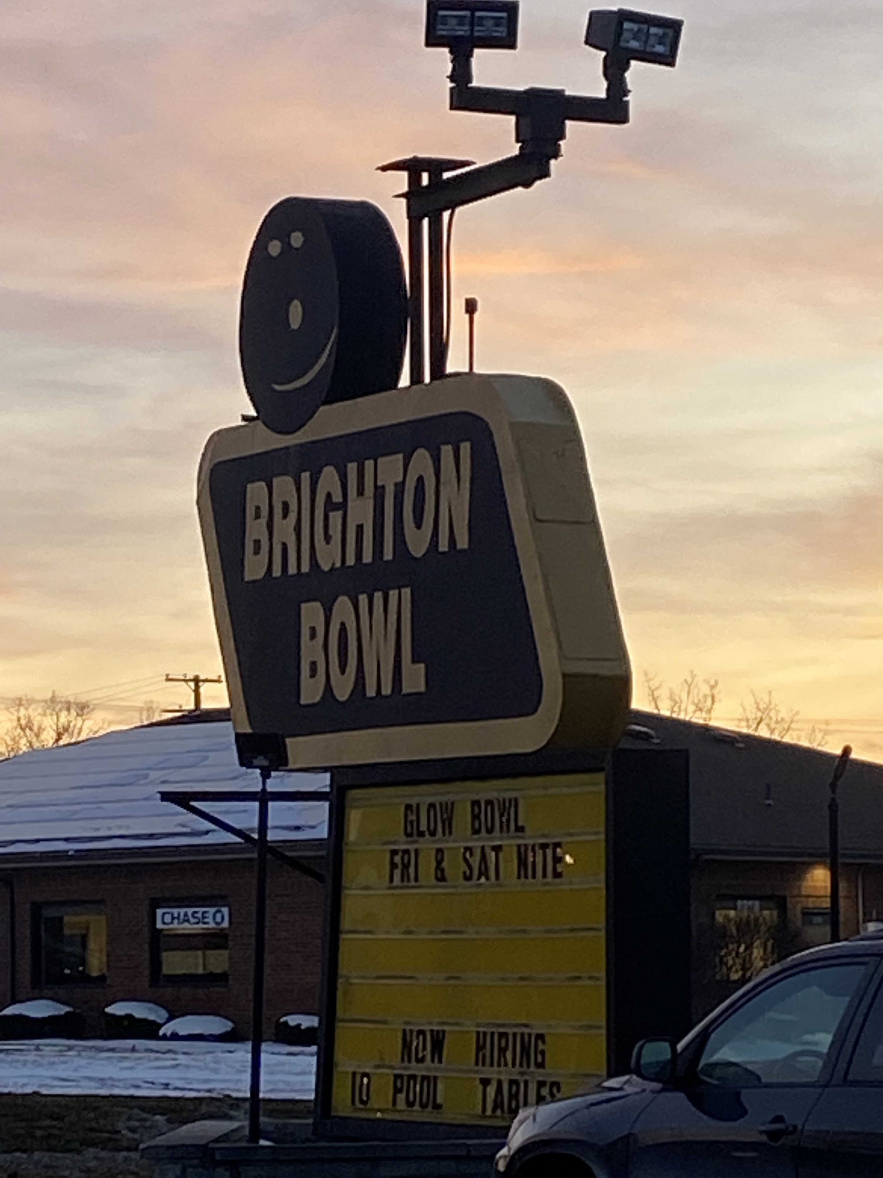 A roadside sign for Brighton Bowl with a bowling ball smiley face icon against a sunset sky.