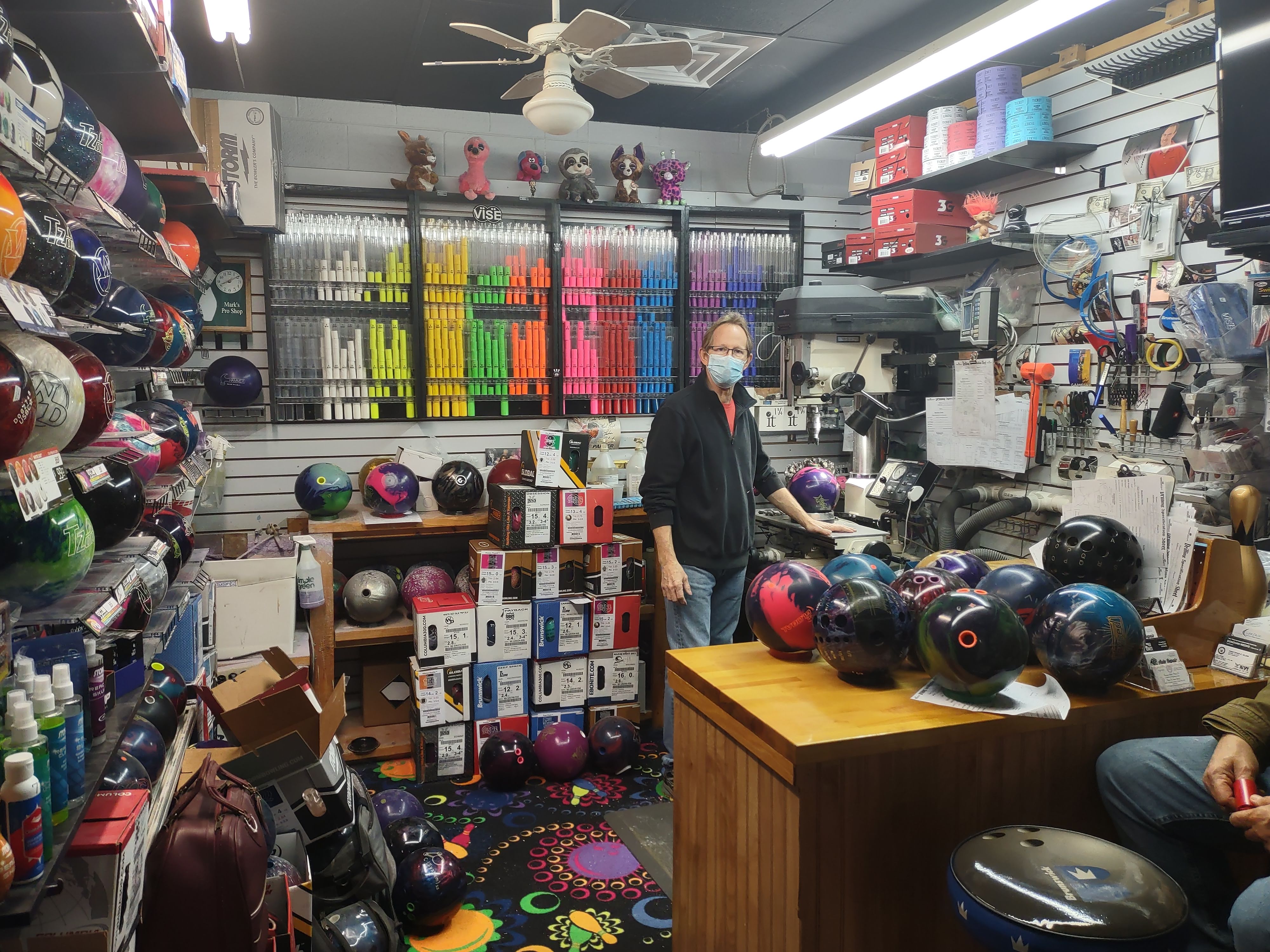 A staff member stands behind a counter in a well-stocked bowling pro shop surrounded by balls and equipment.