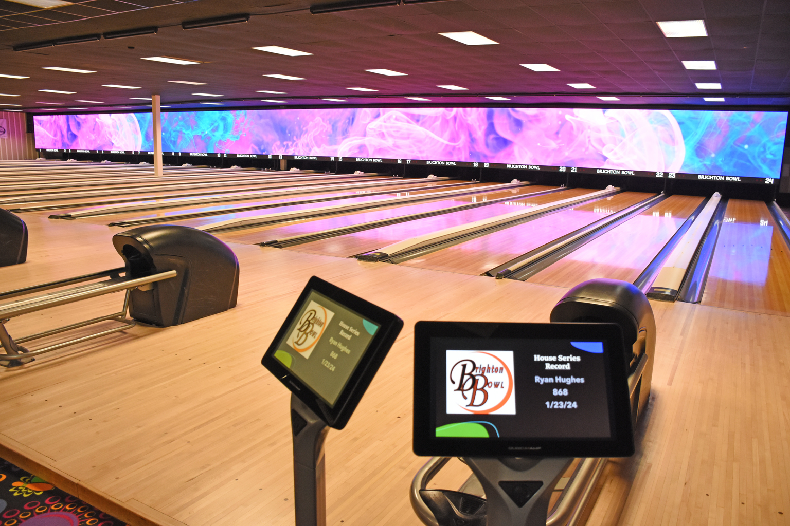 A wide view of modern bowling lanes with vibrant pink and blue digital screens and scoring tablets in the foreground.
