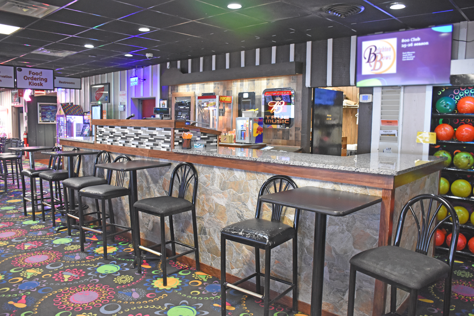A stone-fronted bar with black stools and high-top tables on a colorful patterned carpet inside Brighton Bowl.