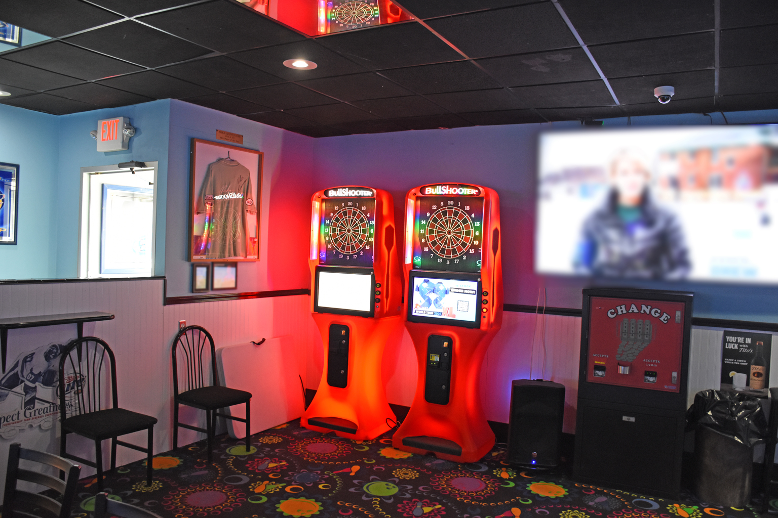 An indoor recreation area featuring two glowing electronic dart boards, a change machine, and seating.