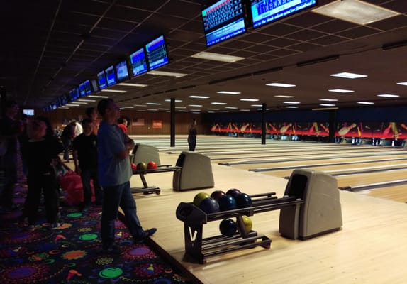 A man stands by a ball return at a bowling alley with several lanes and score monitors visible.