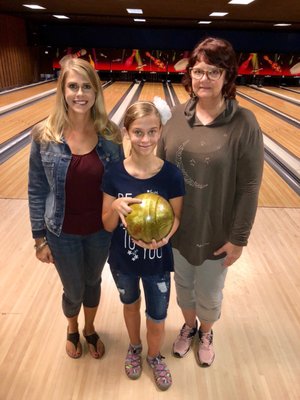 A young girl holding a gold bowling ball stands between two women on a bowling lane.