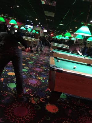 Patrons playing billiards on green tables under bright green lights in a bowling center lounge.