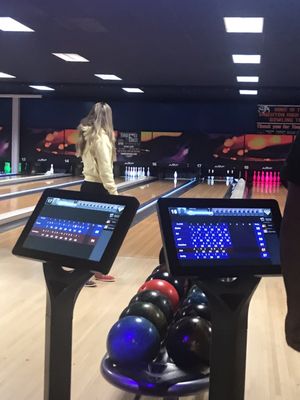 A person stands on a bowling approach behind two digital scoring tablets and a rack of bowling balls.