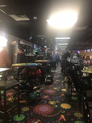 People socializing in a busy bowling center concourse with colorful patterned carpet and a bar in the background.
