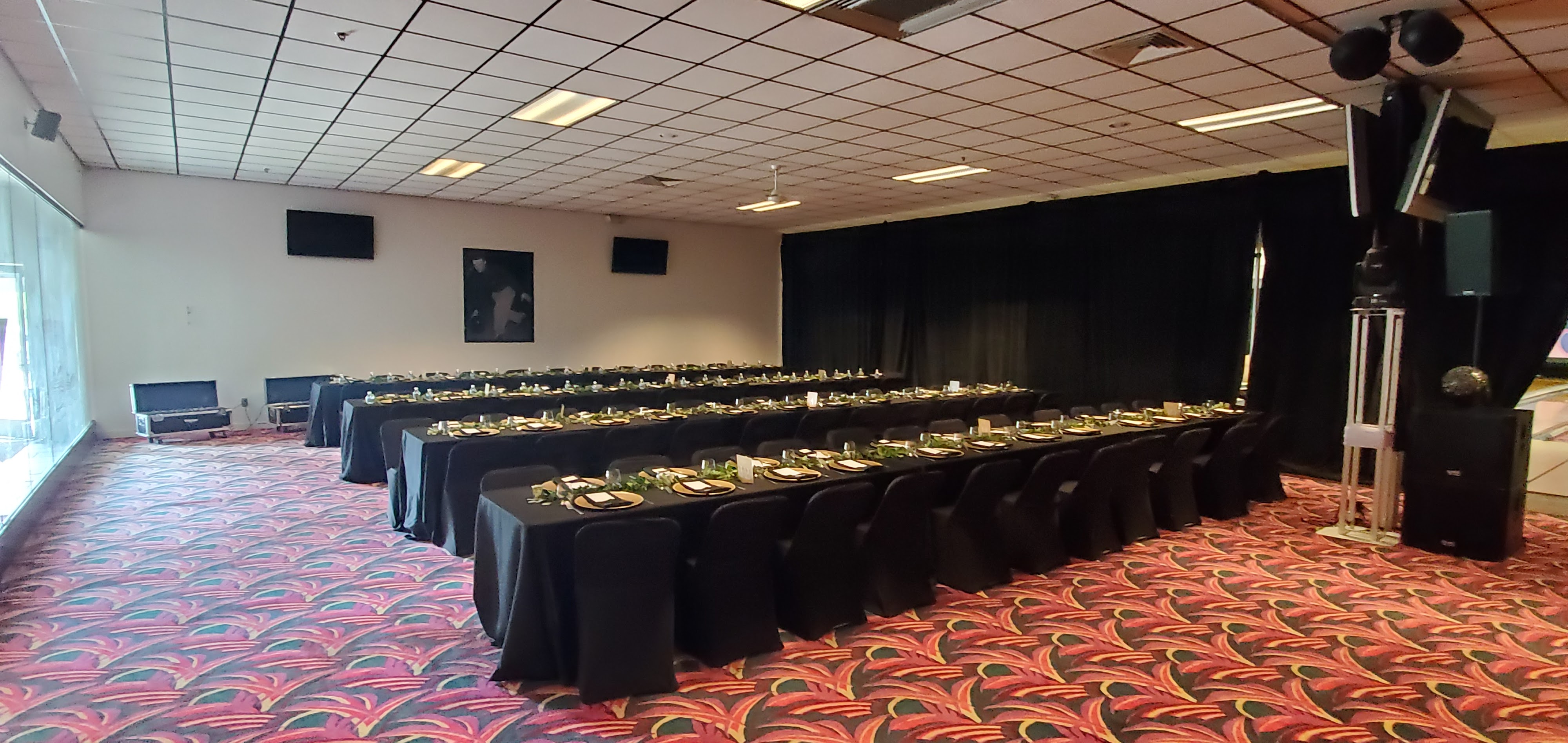 A large event room with several rows of banquet tables set with black linens and gold plates.